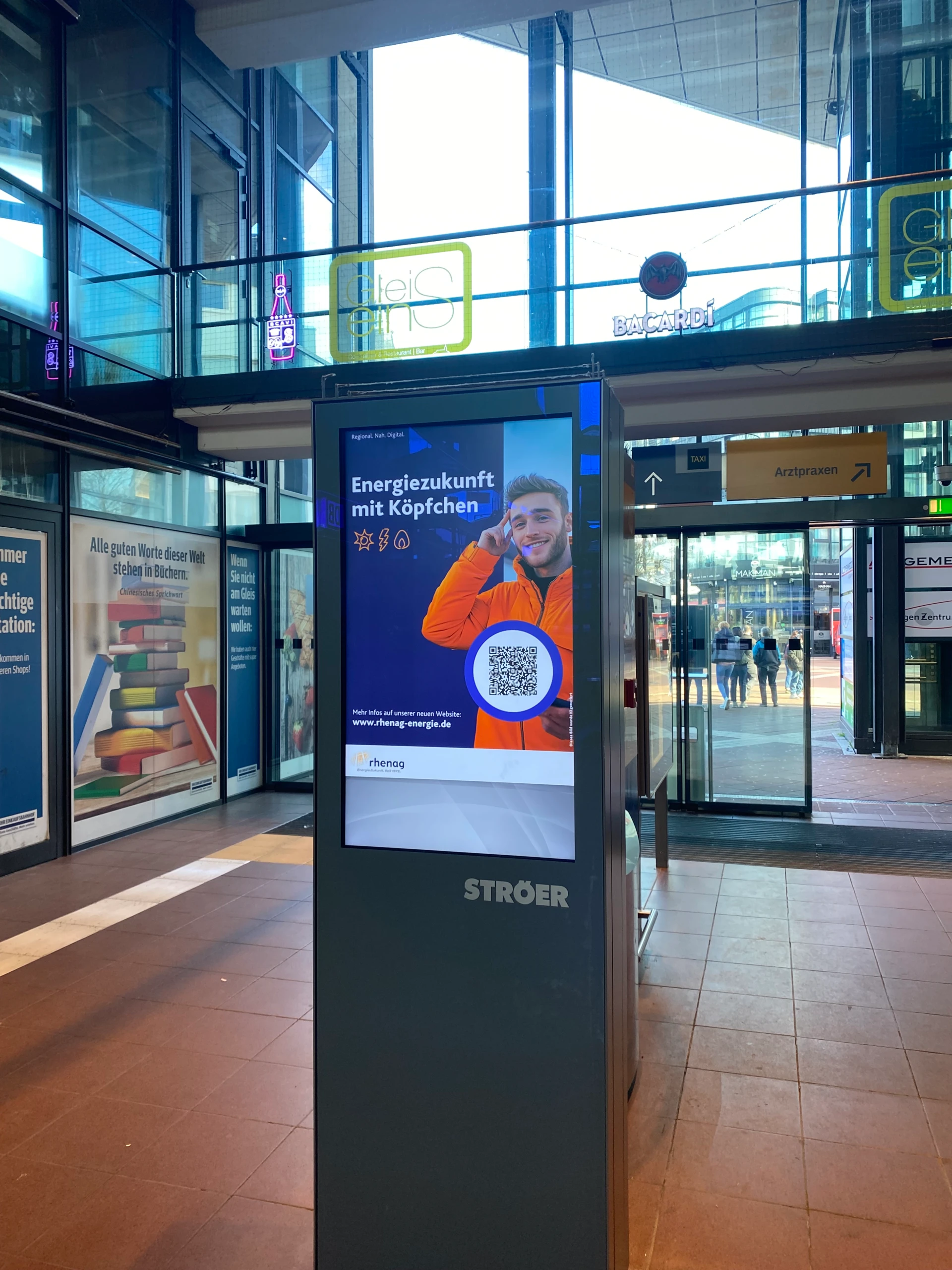 rhenag Kampagne für rhenag-energie.de mit "Energiezukunft mit Köpfchen" als Headline und einem KI-generierten Bild, Icons und QR-Code auf digitaler Werbefläche in einem Bahnhof